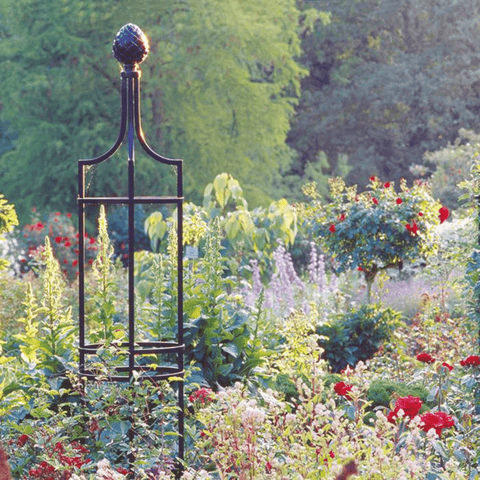 Black metal garden obelisk trellis with rounded top finial in a flower garden bed