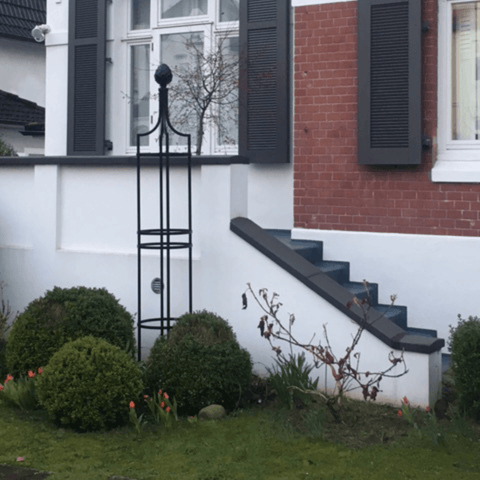 Black metal obelisk trellis with rounded finial and lattice side sections by building wall